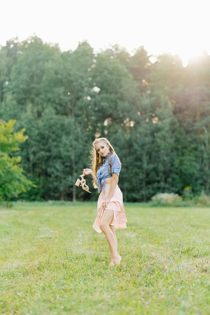 Junge hübsche Frau in einem Sommerkleid geht durch eine Wiese auf grünem Gras Ein junges Mädchen hält Schuhe in ihren Händen, die ihr schönes Bein zeigen Sommererholung im Freien