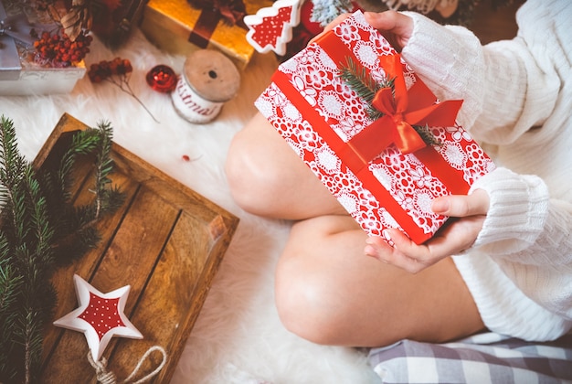 Junge hübsche Frau in warmen, gemütlichen Kleidern sitzt auf dem hellen Holzboden in ihrem hellen Haus in der Nähe des Weihnachtsbaums und hält eine Geschenkbox in ihren Händen.