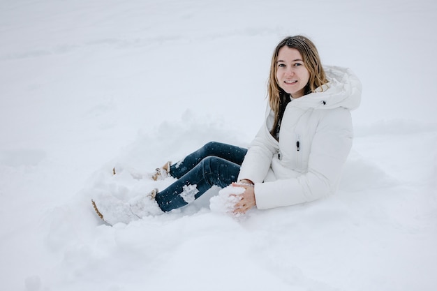 Junge hübsche Frau in Winterkleidung sitzt auf einer Schneeverwehung und lächelt