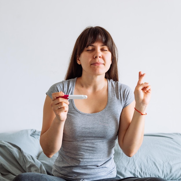 Junge hübsche Frau kreuzt die Finger und wünscht sich einen Baby-Looking-Schwangerschaftstest