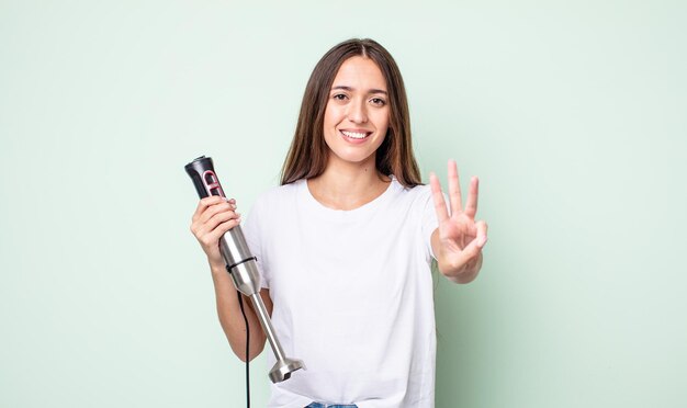 Junge hübsche Frau lächelt und sieht freundlich aus und zeigt das Handmixer-Konzept Nummer drei