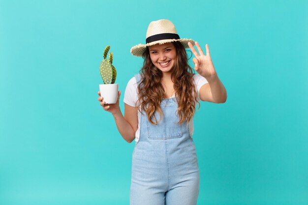 Junge hübsche Frau lächelt und sieht freundlich aus, zeigt Nummer drei und hält einen Kaktus