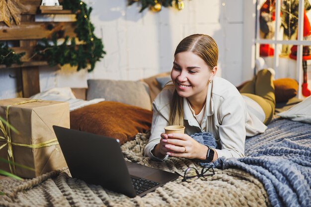 Junge hübsche Frau mit blonden langen Haaren in gemütlicher Kleidung mit Kaffee in den Händen mit Laptop, die Filme anschaut und auf dem Bett im Zimmer mit Weihnachtsbaum zu Hause lächelt Neujahrsferien