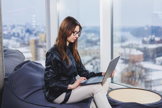 Junge hübsche Frau mit Brille arbeitet an einem Laptop, während sie in einem modernen Arbeitsbereich sitzt Fernarbeit freiberuflich