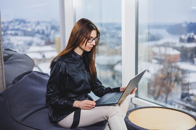 Junge hübsche Frau mit Brille arbeitet an einem Laptop, während sie in einem modernen Arbeitsbereich sitzt Fernarbeit freiberuflich