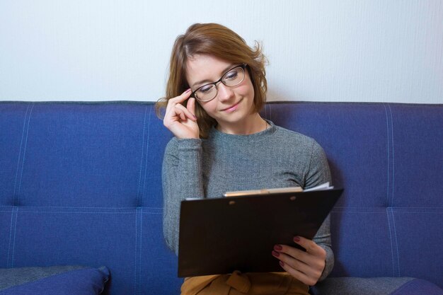 Junge hübsche Frau mit Brille, die auf einem Sofa im Büro sitzt, liest Dokumente