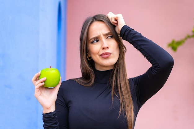 Junge hübsche Frau mit einem Apfel im Freien, die Zweifel hat und einen verwirrten Gesichtsausdruck hat