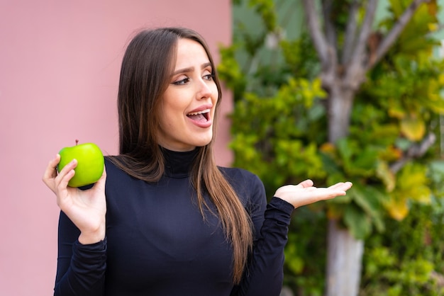 Junge hübsche Frau mit einem Apfel im Freien mit überraschendem Gesichtsausdruck
