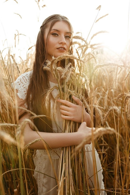 Junge hübsche Frau mit Haufen von Ährchen, die im Sommer bei Sonnenuntergang im Weizenfeld stehen