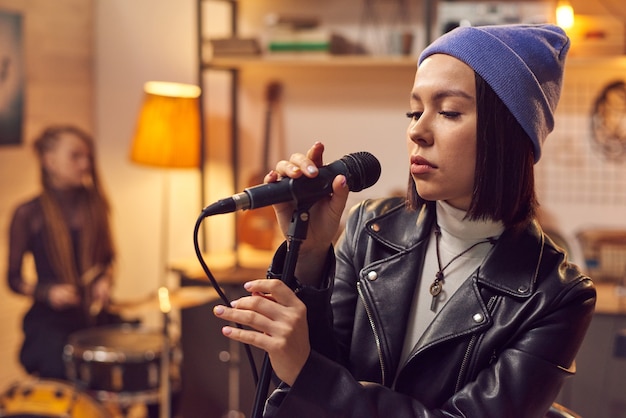 Junge hübsche Frau mit Hut, die während der Probe mit ihrer Gruppe im Studio im Mikrofon singt