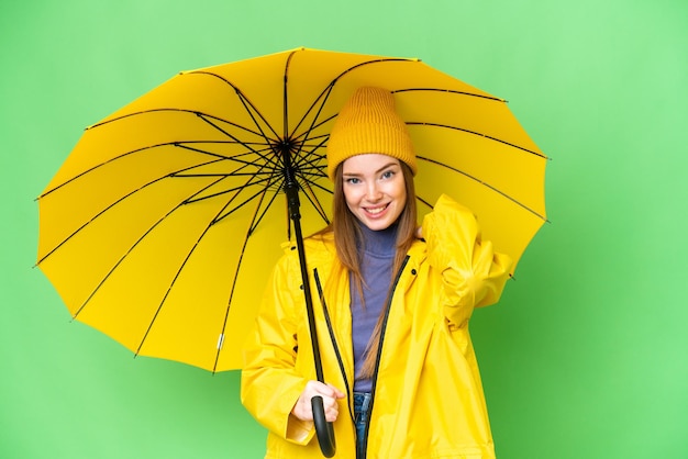 Junge hübsche Frau mit regenfestem Mantel und Regenschirm über isoliertem Chroma-Key-Hintergrund lachend