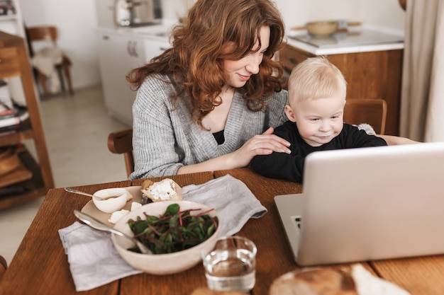 Junge hübsche Frau mit roten Haaren im gestrickten Pullover und im kleinen Sohn, die mit Essen am Tisch sitzen und verträumt Cartoons auf Laptop beobachten