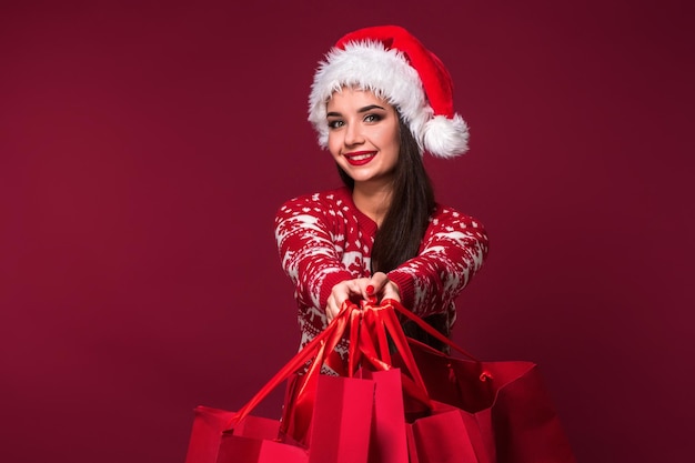 Junge hübsche Frau oder Mädchen mit langen schönen Haaren in Weihnachtsmütze und Neujahrskleid mit rotem Paket auf rotem Studiohintergrund