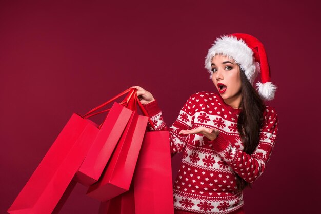 Junge hübsche Frau oder Mädchen mit langen schönen Haaren in Weihnachtsmütze und Neujahrskleid mit rotem Paket auf rotem Studiohintergrund