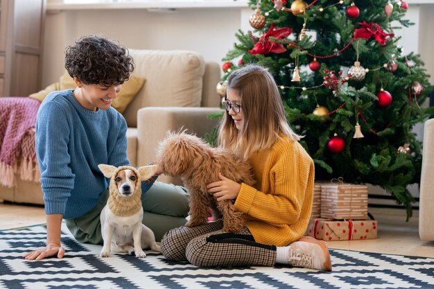 Junge hübsche Frau und ihre Tochter in Freizeitkleidung spielen mit lustigen Hunden, während beide auf dem Boden des Wohnzimmers am Weihnachtsbaum sitzen