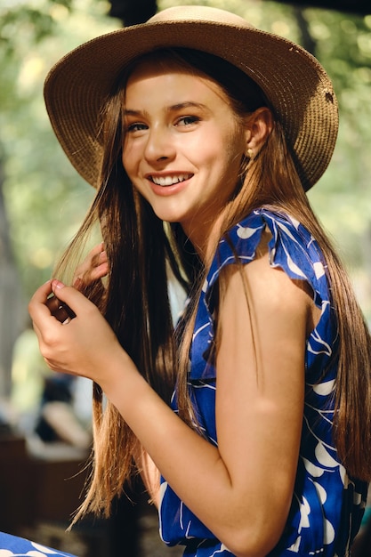 Junge hübsche fröhliche Frau in blauem Kleid und Hut, die glücklich in die Kamera im Stadtpark schaut