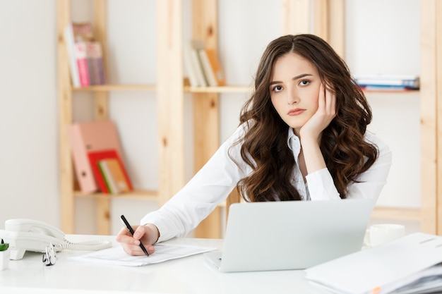 Junge hübsche Geschäftsfrau mit Notizbuch und Dokument im hellen modernen Büro drinnen