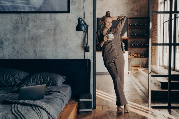 Junge hübsche glückliche Frau, die einen braunen Satinpyjama trägt und eine Kaffeetasse hält, während sie in der Schlafzimmertür steht, nachdem sie morgens zu Hause aufgewacht ist, Frau, die den neuen Tag beginnt