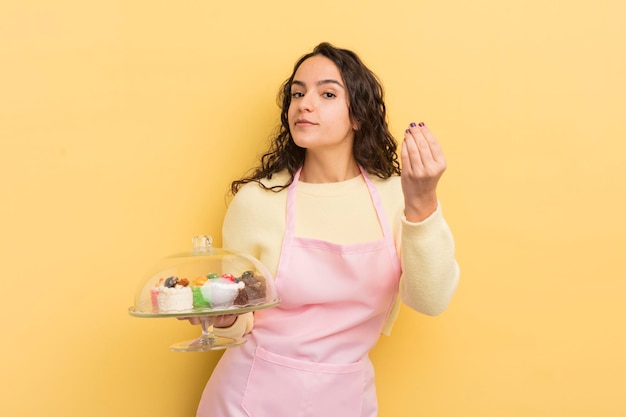 Junge hübsche hispanische frau, die capice oder geldgeste macht, die ihnen sagt, sie sollen das koch- und kuchenkonzept bezahlen