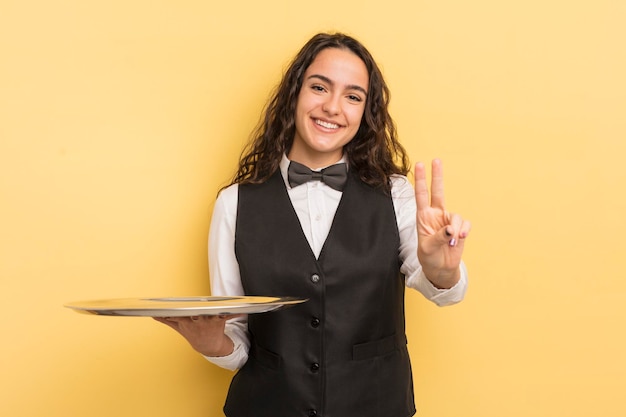 Foto junge hübsche hispanische frau, die lächelt und freundlich aussieht und das kellner- und tablettkonzept nummer zwei zeigt