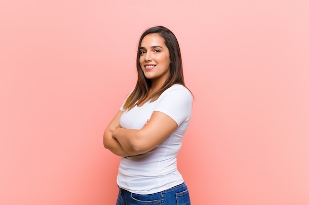 Junge hübsche hispanische Frau, die mit verschränkten Armen und einem glücklichen, selbstbewussten, zufriedenen Ausdruck lächelt, Seitenansicht gegen rosa Wand