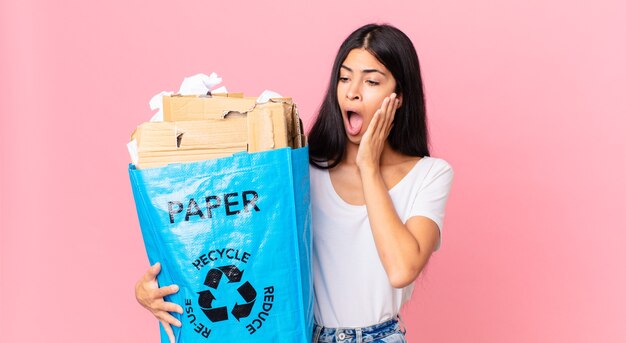 Junge hübsche hispanische Frau, die sich glücklich, aufgeregt und überrascht fühlt und eine Papiertüte zum Recyceln hält