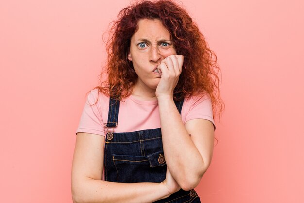 Junge hübsche Ingwer-Rothaarigefrau, die einen Jeanslatzhose beißt die Fingernägel, nervös und sehr besorgt trägt.