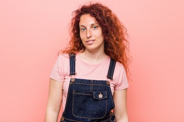 Junge hübsche Ingwer-Rothaarigefrau, die einen Jeanslatzhose glücklich, lächelnd und nett trägt.