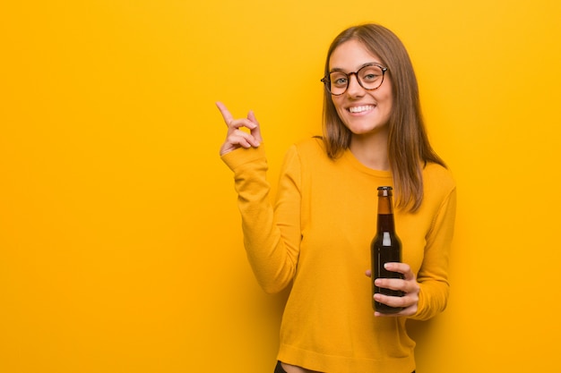 Junge hübsche kaukasische Frau, die auf die Seite mit dem Finger zeigt. Sie hält ein Bier.