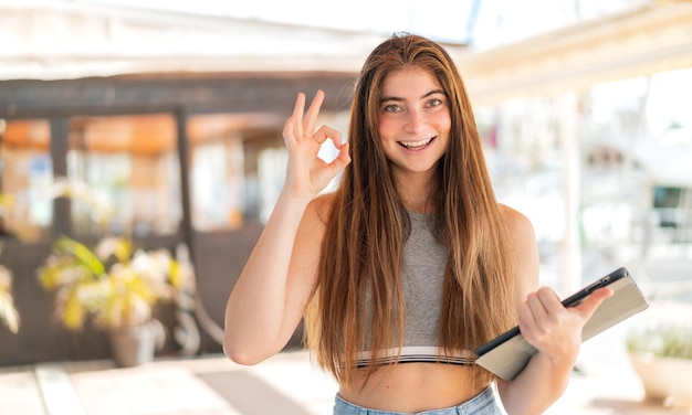 Junge hübsche kaukasische Frau hält im Freien ein Tablet und zeigt mit den Fingern das OK-Zeichen