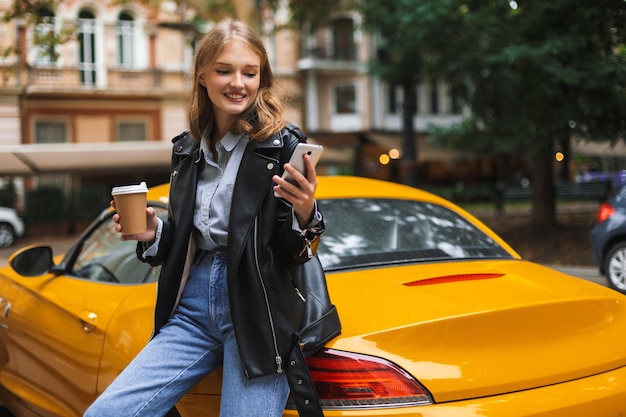 Junge hübsche lächelnde Frau in Lederjacke, die sich auf einen gelben Sportwagen stützt und eine Tasse Kaffee in der Hand hält, während sie glücklich mit dem Handy auf der Straße der Stadt unterwegs ist