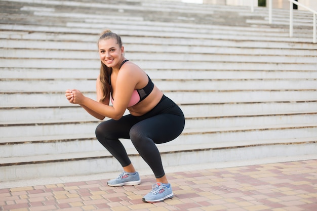 Junge hübsche lächelnde Plusgrößenfrau im sportlichen Oberteil und in den Leggings, die Sport mit Treppen tun