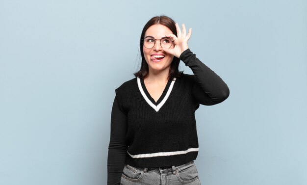 Junge hübsche lässige Frau, die glücklich mit lustigem Gesicht lächelt, scherzt und durch Guckloch schaut, Geheimnisse ausspioniert