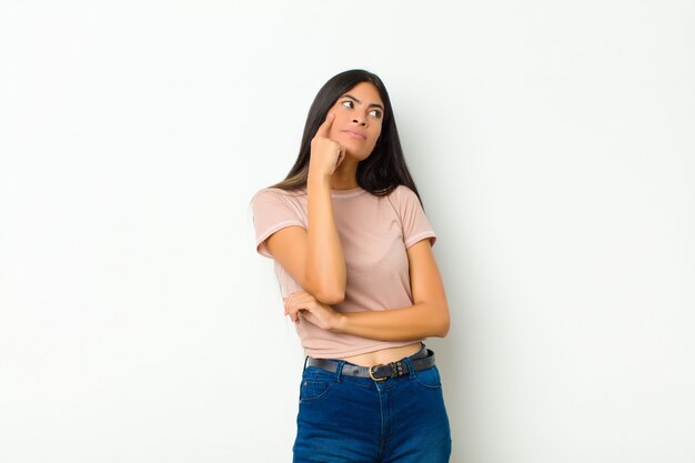 Junge hübsche lateinamerikanische Frau mit einem konzentrierten Blick, der sich mit einem zweifelhaften Ausdruck wundert und zur flachen Wand zur Seite schaut