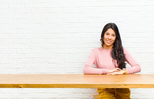 Junge hübsche lateinische Frau, die freundlich und zufällig mit einem positiven, glücklichen, überzeugten und entspannten Ausdruck sitzt vor einer Tabelle lächelt