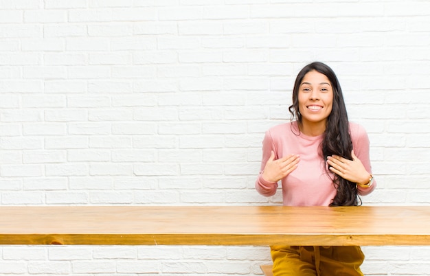 Junge hübsche lateinische Frau, die glücklich, überrascht, stolz und aufgeregt schaut und auf das Selbst zeigt, das vor einer Tabelle sitzt