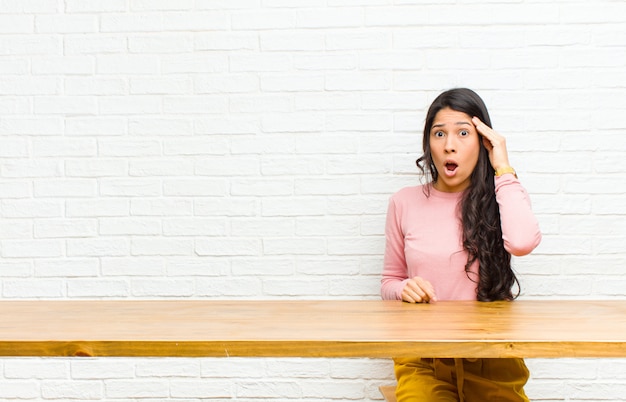 Junge hübsche lateinische Frau, die überrascht, mit offenem Mund, entsetzt, einen neuen Gedanken, eine Idee verwirklichend oder vor einem Tisch sitzend schaut