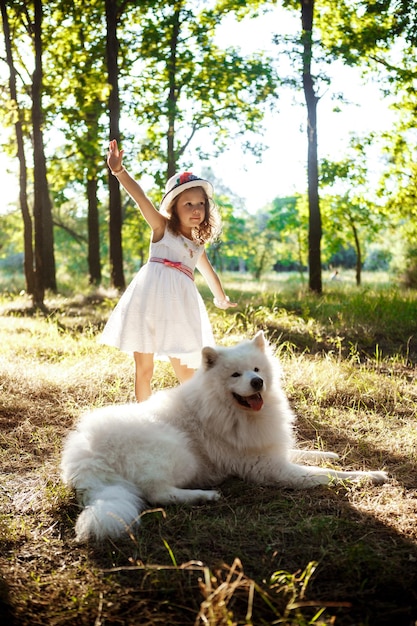 Junge hübsche Mädchen in Kleid und Hut zu Fuß, spielen mit weißem Hund im Park bei Sonnenuntergang.