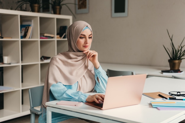 Junge hübsche moderne muslimische Frau im Hijab, die am Laptop im Büroraum arbeitet, Bildung online