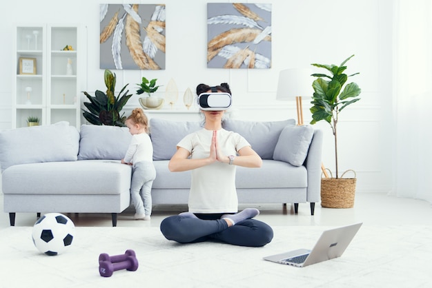 Junge hübsche Mutter meditiert in Lotus Yoga Position mit Virtual-Reality-Brille, während ihre Tochter Cartoons zu Hause auf Hintergrund sieht.