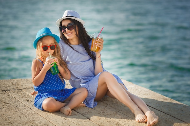 Junge hübsche Mutter und ihre kleine Tochter am Strand haben Spaß. Mädchen trinken Limonade.