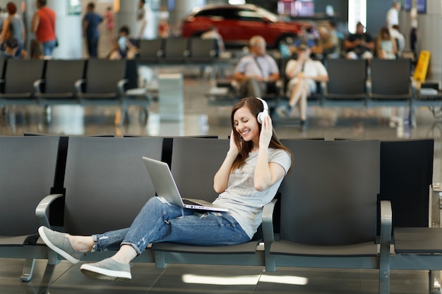 Junge hübsche Reisende Touristenfrau mit Kopfhörern, die Musik hören, die am Laptop arbeitet, warten in der Lobbyhalle am internationalen Flughafen?