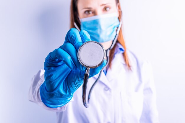 Junge hübsche Ärztin mit Handschuhen, Maske und Kittel, die ein Stethoskop vor weißem Hintergrund hält