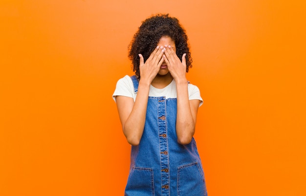 Junge hübsche schwarze Frau, die traurig, frustriert, nervös und depressiv fühlt, Gesicht mit beiden Händen bedeckt, gegen orange Wand weint