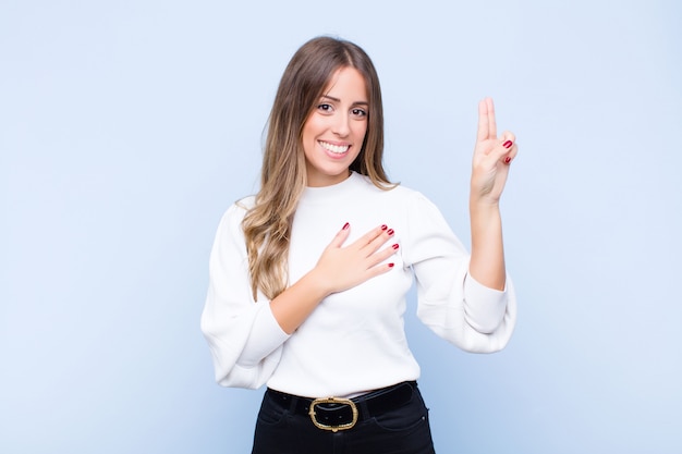 Junge hübsche spanische Frau, die glücklich, zuversichtlich und vertrauenswürdig aussieht, lächelt und Siegeszeichen zeigt, mit einer positiven Haltung gegen blaue Wand