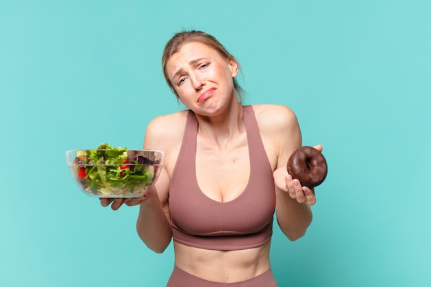 Junge hübsche Sportfrau traurige Ausdruckshand, die ein Sandwich und einen Donut hält