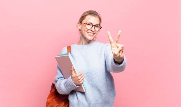 Junge hübsche Studentin, die lächelt und freundlich aussieht, die Nummer zwei oder die zweite mit der Hand nach vorne zeigt, herunterzählt