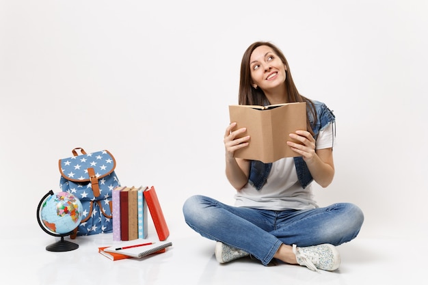 Junge hübsche Studentin in Denim-Kleidung, die träumt, ein Buch zu halten, das in der Nähe des Globus-Rucksack-Schulbücher sitzt