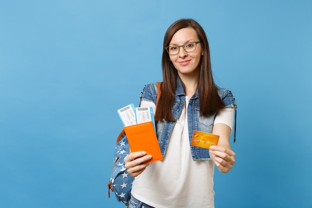 Junge hübsche Studentin in Gläsern mit Rucksack mit Reisepass, Bordkarten, Kreditkarte auf blauem Hintergrund isoliert. Ausbildung an einer Hochschule im Ausland. Flugreise-Flugkonzept.