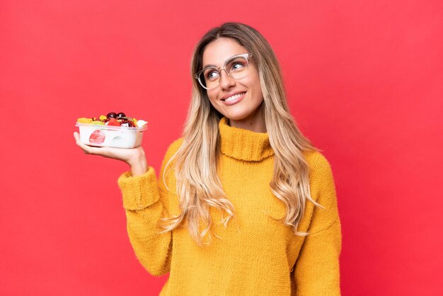 Junge hübsche uruguayische Frau, die eine Obstschale isoliert auf rotem Hintergrund hält und eine Idee denkt, während sie nach oben schaut